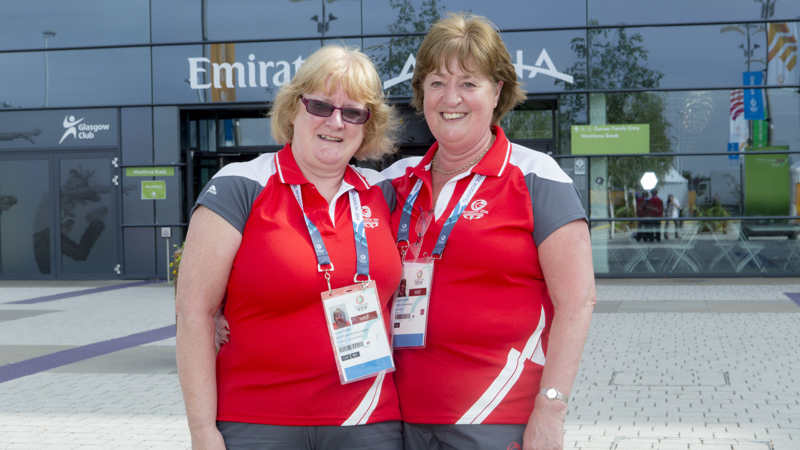 Two volunteers wearing the red Clydesider uniform during the Glasgow 2014 Commonwealth Games. They are smiling and standing outside the Emirates Arena.