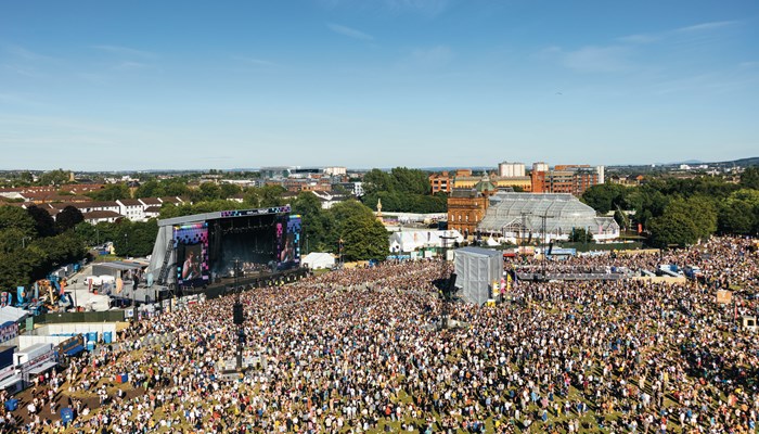 An aerial view of the main stage of Trnsmt festival in Glasgow Green. The stage is lit up and there is a huge crowd shown on a sunny day. Surrounding Glasgow buildings and housing can be seen in the background.