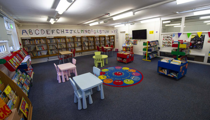 A view of the spacious children's area. There is an alphabet above a row of bookshelves, colourful tables and chairs, books and a rug with faces in different colours.