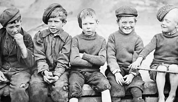 A black and white photo of a group of 5 children sitting on a wall, bare footed, wearing rags, hats and one had a stick.