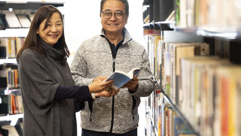 two people facing the camera and smiling, standing beside book shelves holding a book. 