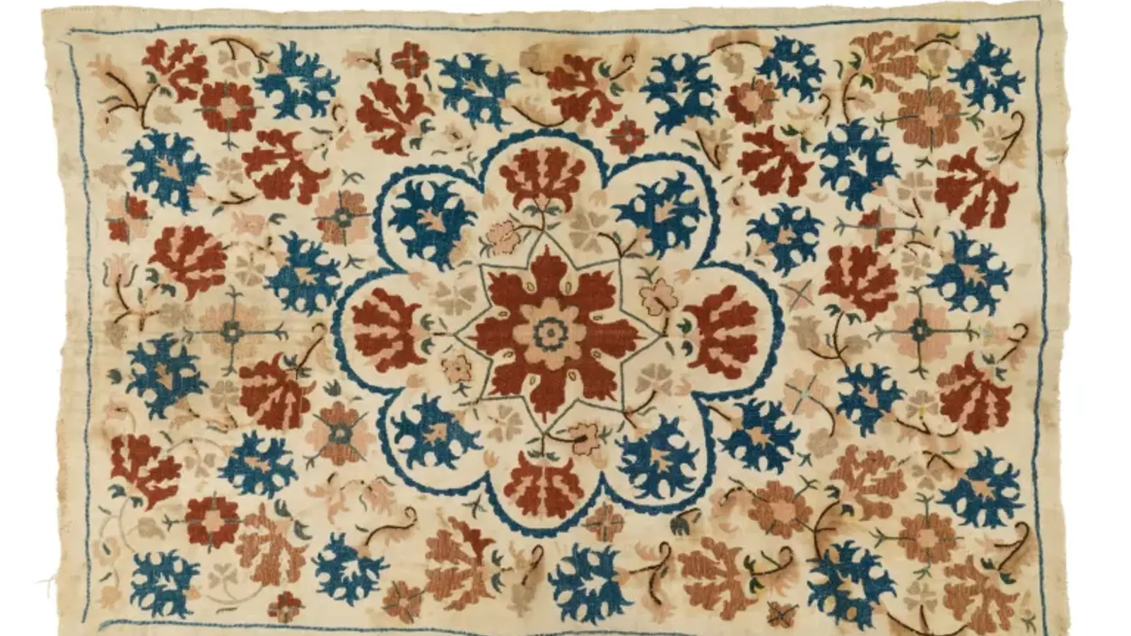 an image of an embroidered rug with red and blue patterns on a cream base.