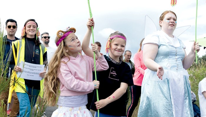 A mix of people dressed up and walking outdoors as part of a parade. One person is wearing a long silver dress with wings and carrying a flower mobile. In the foreground two children are carrying a home made banner with a green curved leaf.