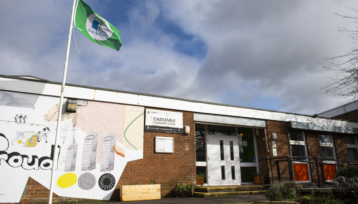 exterior shot of a community centre made of brown brick. there is a sign that says castlemilk community centre in white and black. there is a green flag on a flagpole outside the venue. there is a mural on the side of the build with the work proud printed beside the entrance