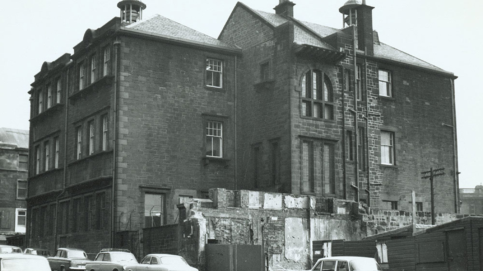 A black and white photo of a large sandstone building, with several chimneys. Cars are parked along the road.