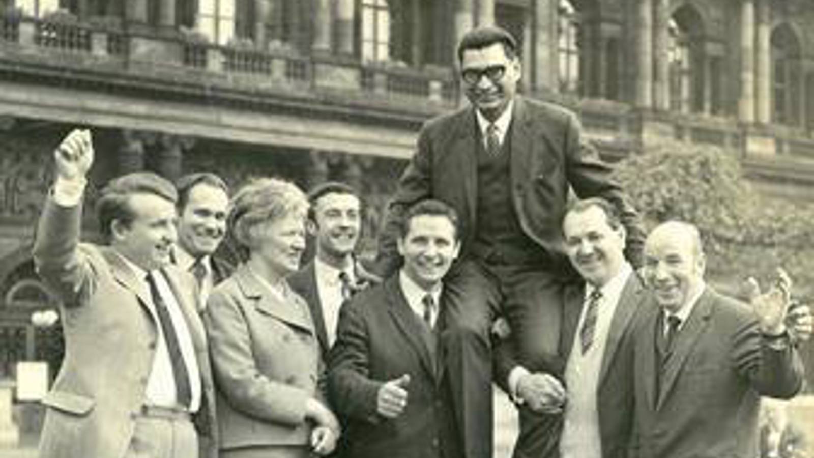 One person wearing glasses smiling while sitting on the shoulders of two people, with another 5 people celebrating with them by waving hands in the air and smiling. There is a large building and shrubs in the background.