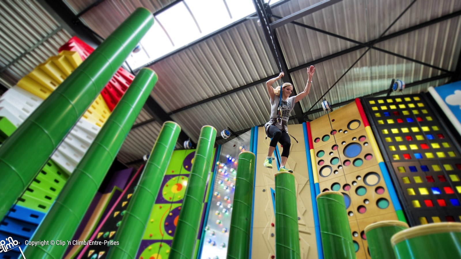 A person with a harness on leaping from one high green soft pillar to another at height. Multicoloured climbing wall features are in the background.