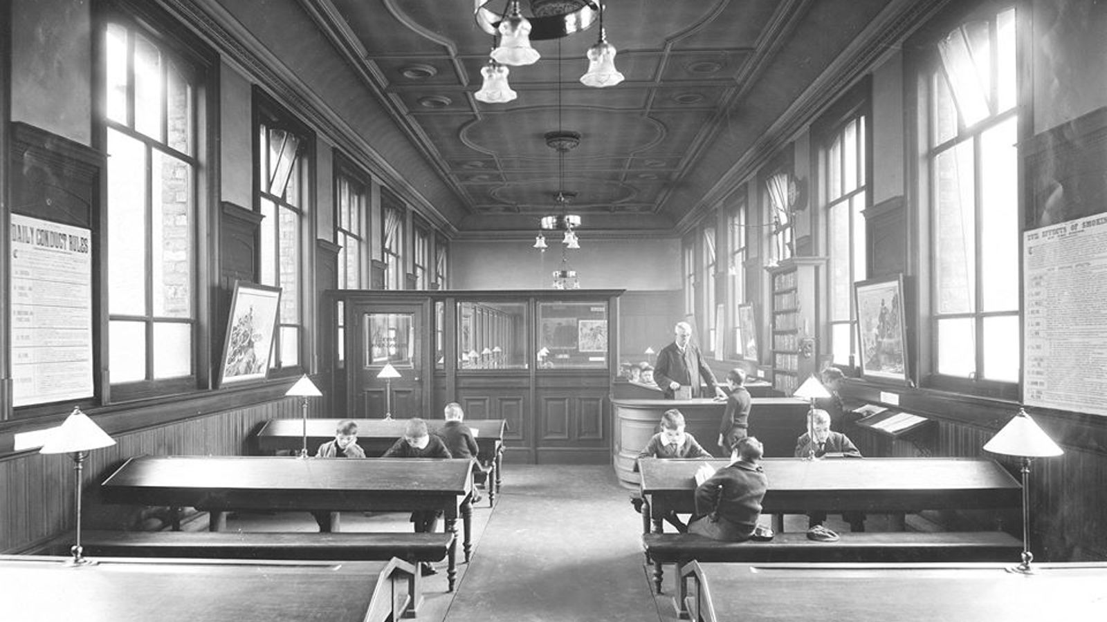 A black and white photograph of inside Bridgeton Library. There are three rows of tables with lamps, with 8 children reading and a librarian at a curved wooden desk.