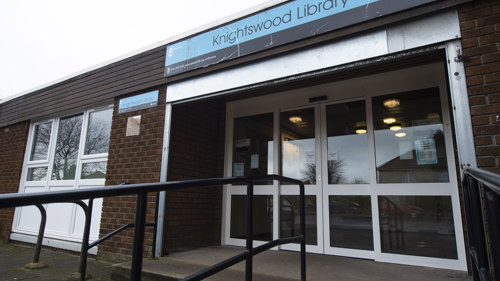 Double automatic entrance doors, to red brick building, blue sign above the door reads Knightswood Library. 