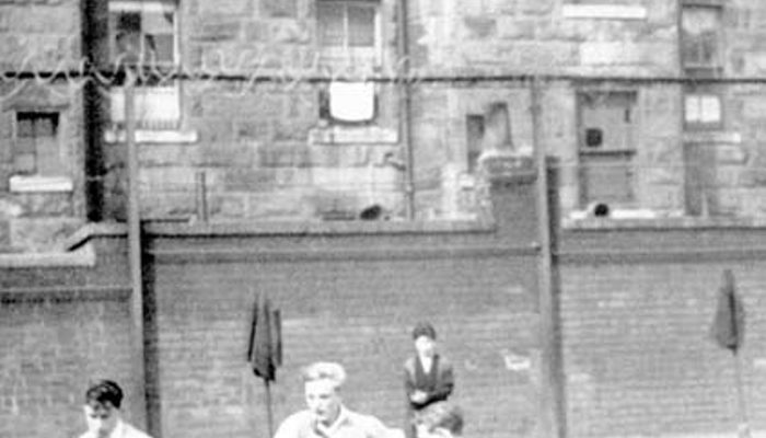 Four young people playing football in a playground secured with barbed wire with large buildings overhead