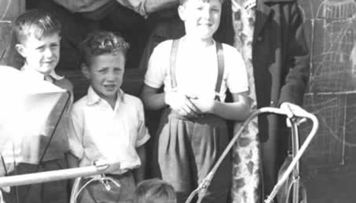 Black and white photo showing an older women standing with 4 children, one of them in a pram with a man leaning out a window behind them