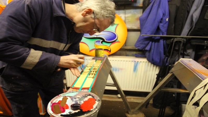 A person with glasses on painting letters on a sign using a paint brush and palette. There is artwork in the background.