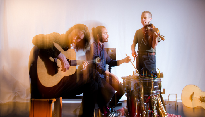 Three musicians from the band Coras Trio, one is sitting playing an acoustic guitar, another the drums and the third is standing up playing the violin.