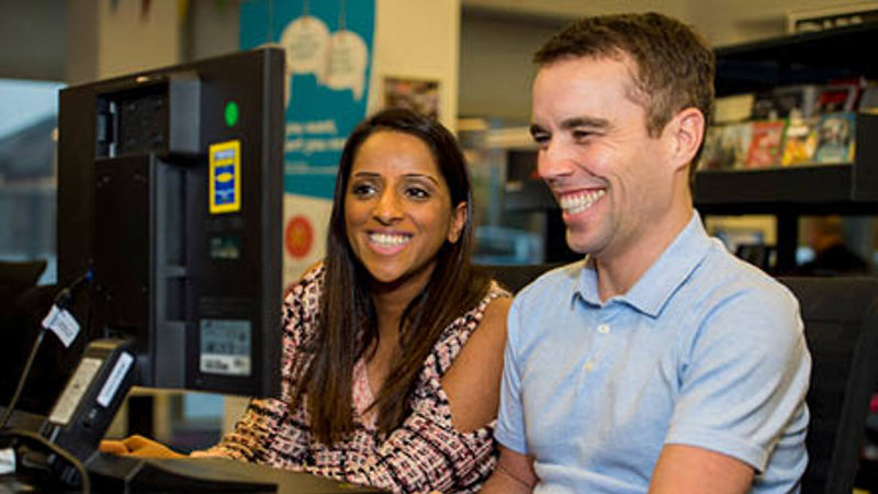 Two people are sitting down in a library smiling and looking at a computer screen together