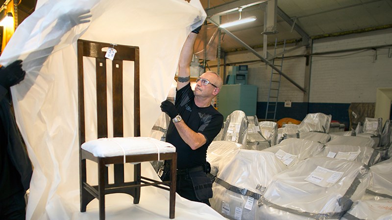 Two people unwrapping plastic from a restored Charles Rennie Mackintosh chair.  The image is taken in a warehouse with a number of other items wrapped in plastic in the background.