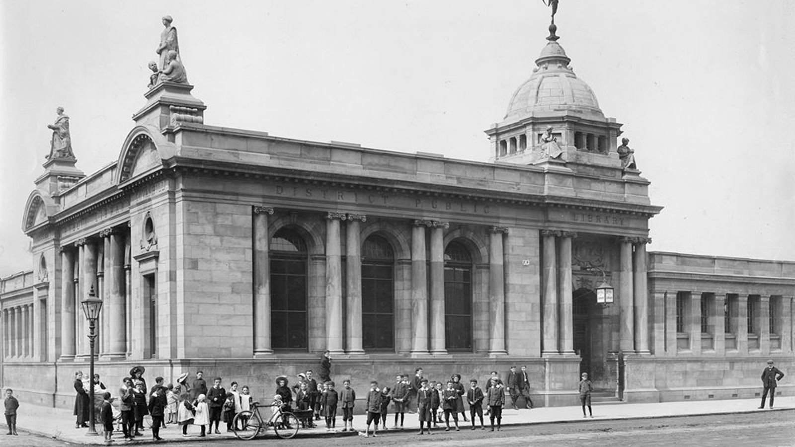 A black and white photograph of Govanhill Library which architecture includes pillars, arches and statues. Many people are on the street outside including adults, children and a bike.