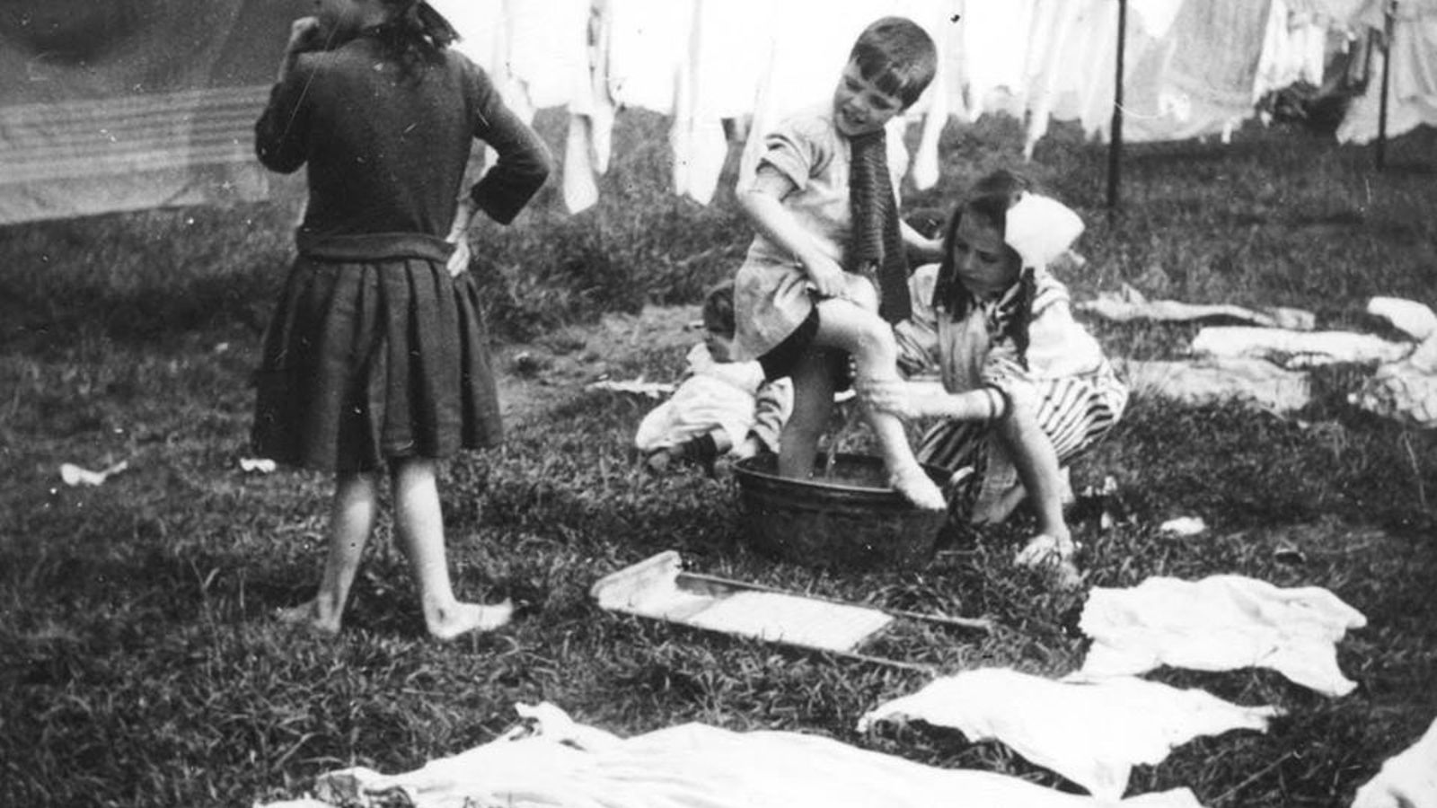 Black and white photograph of children washing in a garden. One child is standing in a tub getting their legs washed by a sligtly older child. Two younger children are sitting on the ground and an older child is standing up looking the other way. Lots of washing it drying on a line and the grass.