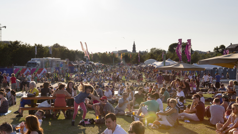 A large crowd of people sitting in the Glasgow Green fan zone during the 2014 Commonwealth Games on a sunny evening.