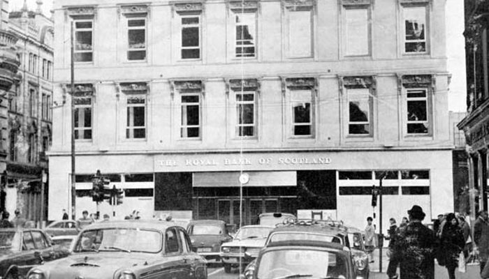 A photo of a three storey building of a bank with old fashioned cards parked outside and people walking in and out of the bank.