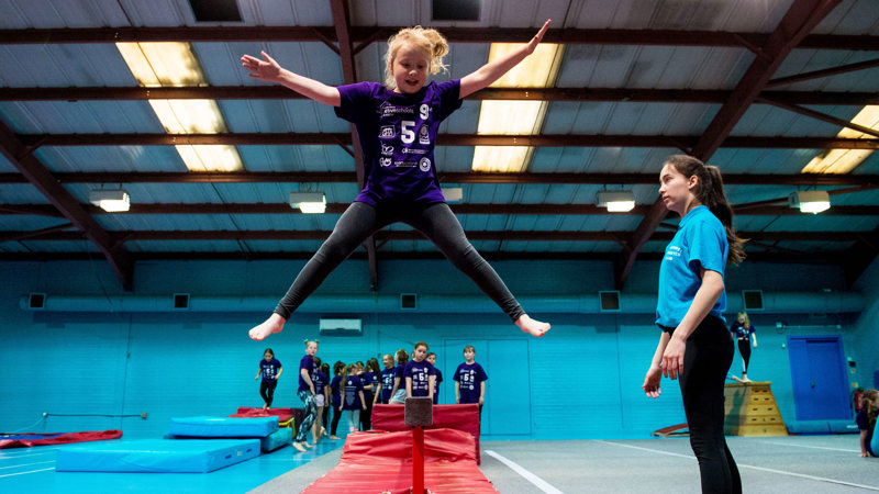 A young person in mid-air while doing a star jump off a balance beam during a gymnastics session as a coach watches her. Other young people are queued up behind her waiting for their shot.