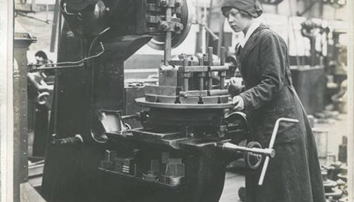 A black and white photo of an employee operating on a large slotting machine, wearing a long dark uniform with white collar and hat.