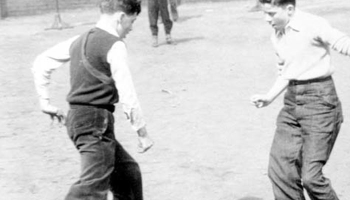 Black and white photo of four young people playing football in a playground with large buildings overhead