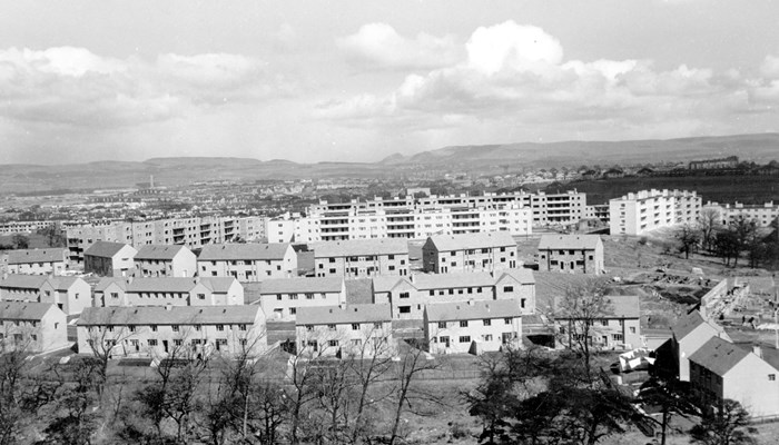 Black and white photograph of housing in Glasgow, with light coloured buildings including houses and flats with trees at the foreground and clouds overhead.