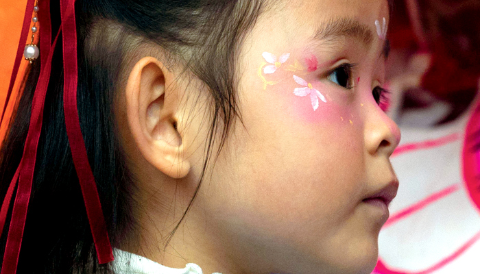 A side view of a child wearing a pale blue dress. They have their face painted with flowers and their hair is tied in a red ribbon with a dangling jewel.