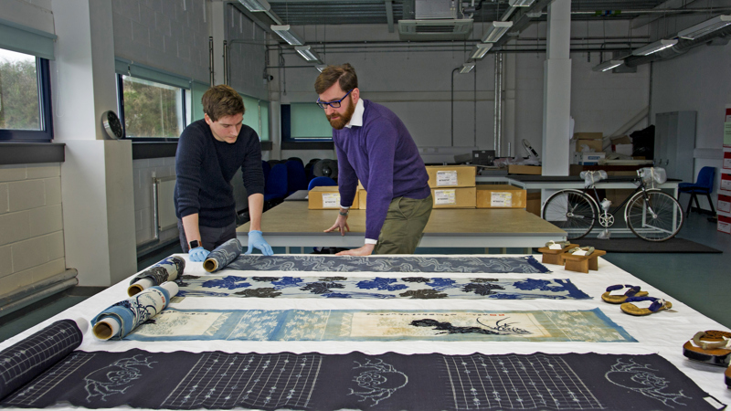 Two curators at work in the Research Room at GMRC viewing textile objects laid flat on a table