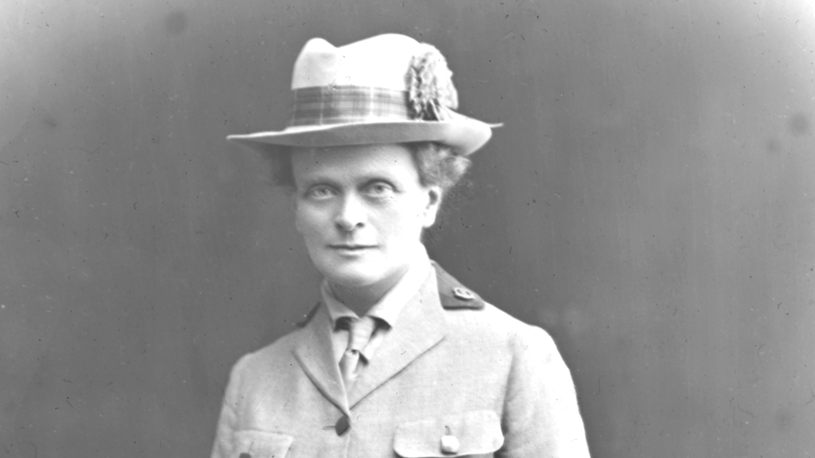 A black and white photo of Elsie Maud in a light coloured uniform, including a tie and a hat, grasping a pair of gloves in her left hand.