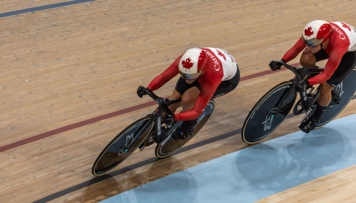 Two riders from the Canadian cycling team, one is in front of the other, during a track race at the Sir Chris Hoy Velodrome during the 2023 UCI Cycling World Championships.