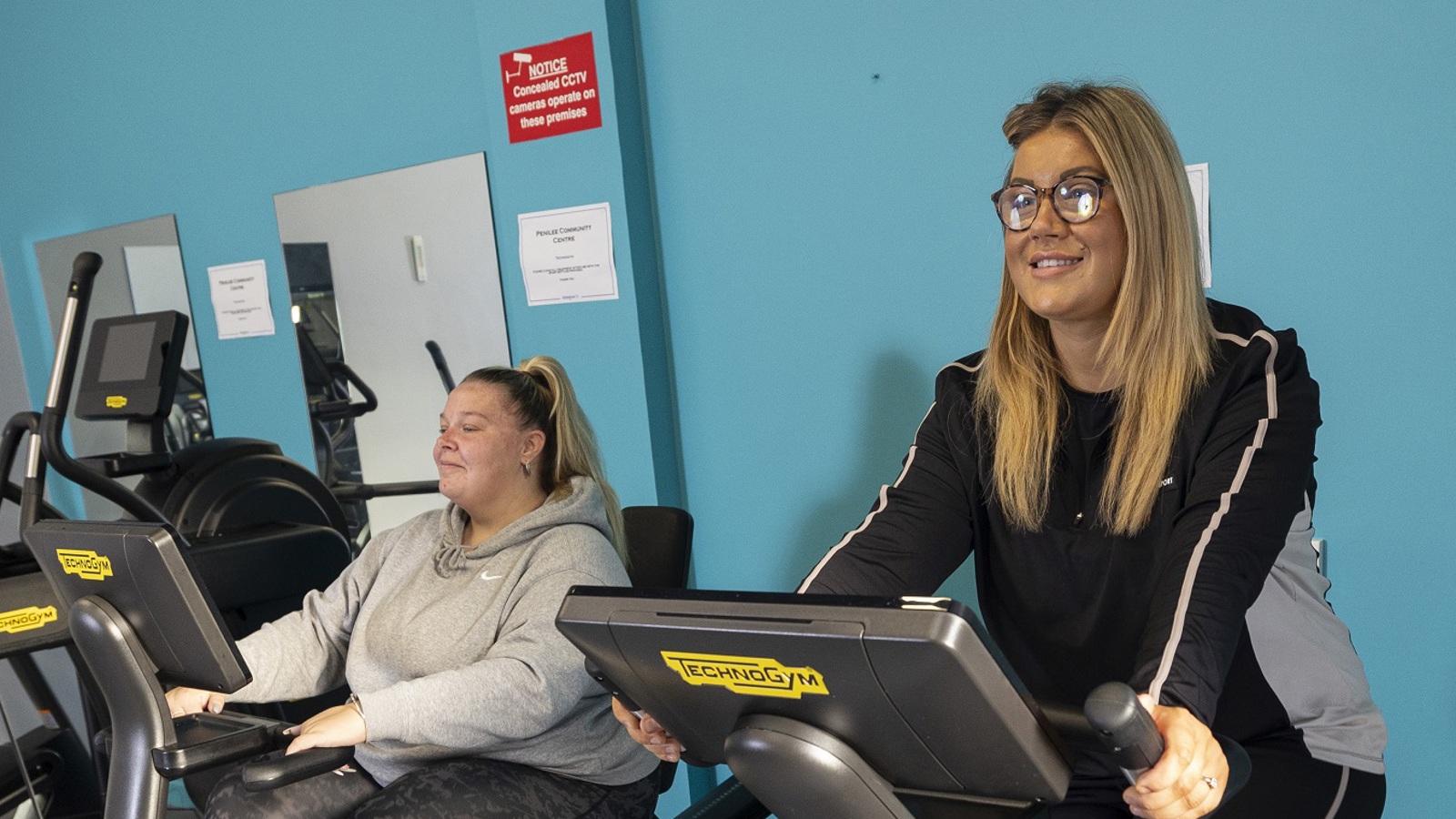 Two people sitting on static bikes in an indoor gym space in a community centre