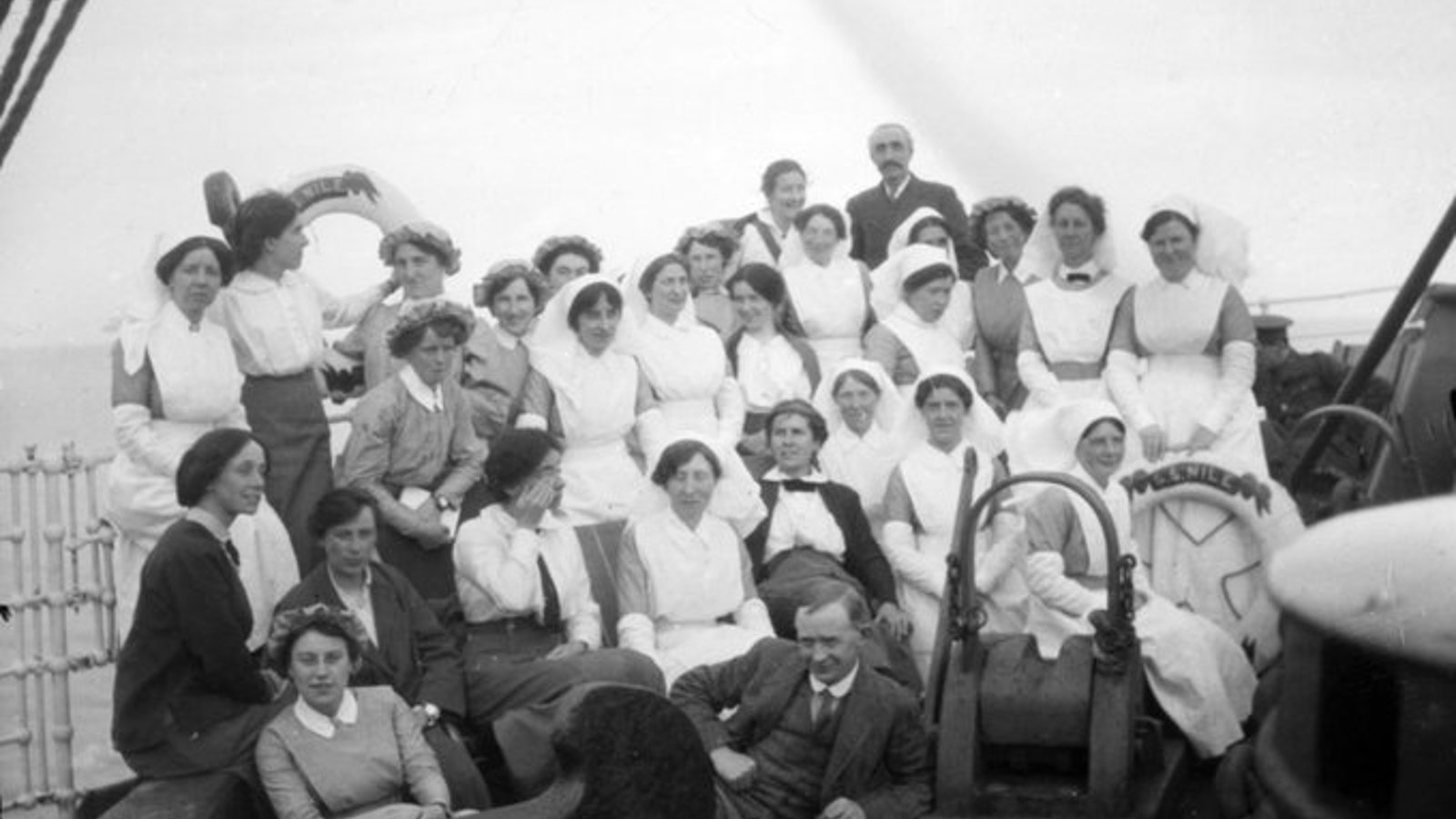 A black and white photograph of a group of people from the Scottish Women's Hospital on a ship wearing white uniforms and dark suits.