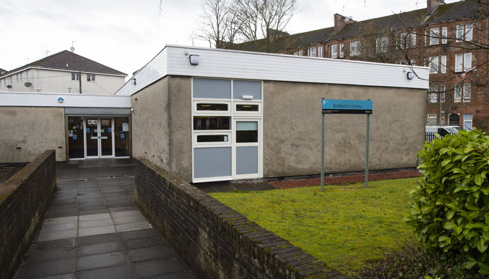 Exterior of Bailliston Library which is a flat roof building with white double doors. There is a small patch of grass outside the library and there are red sandstone tenement flats in the background.