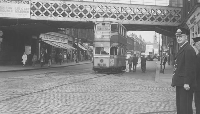 A black and white photo of a tram driving under a bridge, coming round a corner, on a cobbled road, lots of people walking on the pavement.