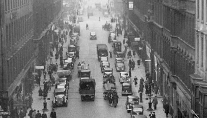 A black and white photo of a busy street with horse and carriages, pedestrians, many sandstone tall shop buildings and many shop fronts.