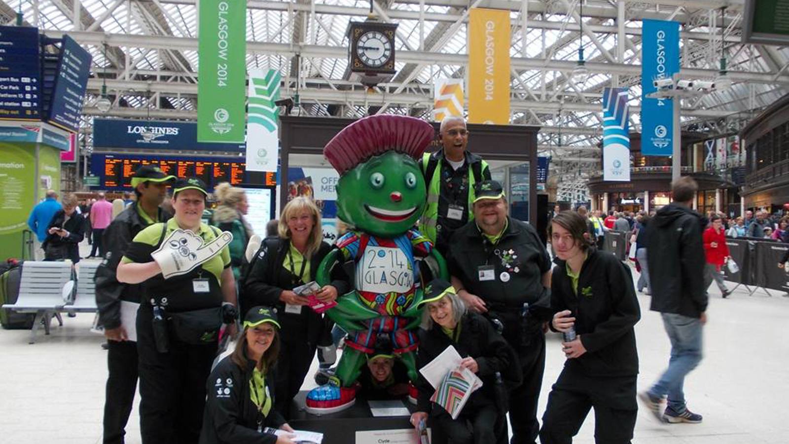 Host City volunteers in front of a Clyde the mascot statue in Glasgow Central Train Station during the Glasgow 2014 Commonwealth Games. The station is busy and has lots of Glasgow 2014 signs hanging from the roof.