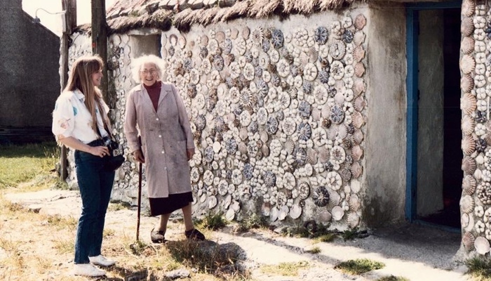 Flora is standing outside a cottage decorated with shells on the outside. She is smiling and leaning on a walking stick. Beside her is another person with a camera around their neck.