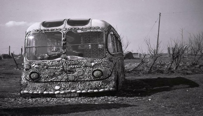 The front of Flora's shell bus. The bus is in a field and apart from the windows the front is covered in shells.