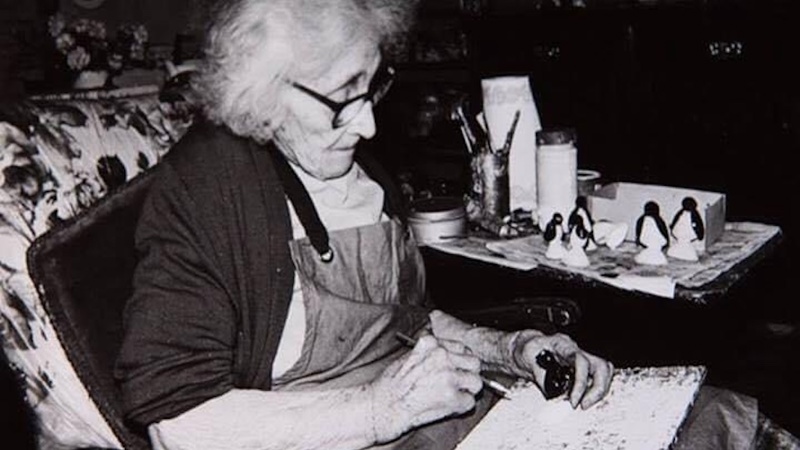 Flora is shown sitting in a chair. She has glasses on and is wearing a dark cardigan and has an apron on. Flora is painting and there is a table to her left with drawing materials.