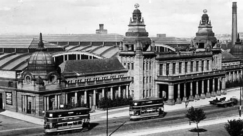 Old black and white photograph of the outside of the Kelvin Hall in Glasgow. There are trams pictured in the front of the building. The building has three towers to the front.
