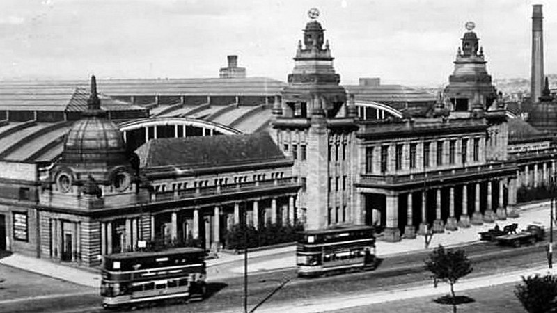 Old black and white photograph of the outside of the Kelvin Hall in Glasgow. There are trams pictured in the front of the building. The building has three towers to the front.