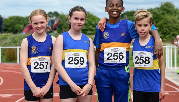 Four young athletes, representing different genders and ethnicities, standing side by side on a running track. They are all wearing the blue and white club colours of Shettleston Harriers.