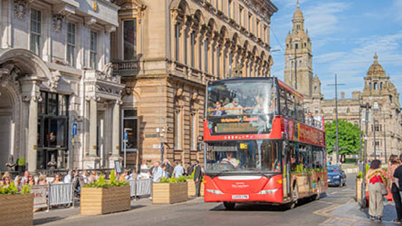 A red double decker bus is driving on a road, people are sitting at tables enjoying food and drink, there is sandstone buildings and blue skies behind the bus