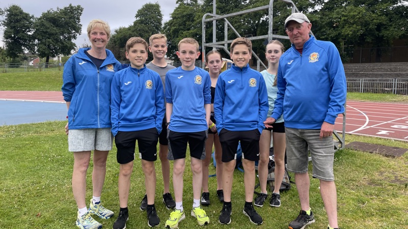 Two adult coaches from Shettleston Harriers with a group of young athletes, who are a mix of genders, at Crownpoint athletics track in Glasgow.