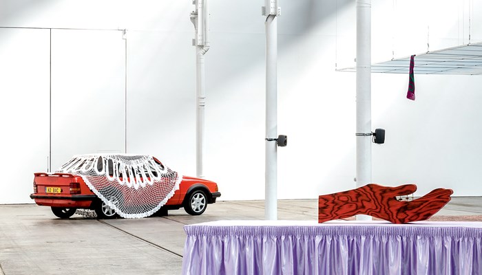 Art installation by Jasleen Kaur. The exhibition, Alter Altar shows a red car partly draped by a large white crocheted shawl/covering. There is a model of an upturned red hand with black markings in the foreground. The exhibits are in Tramway in an industrial setting with pillars.