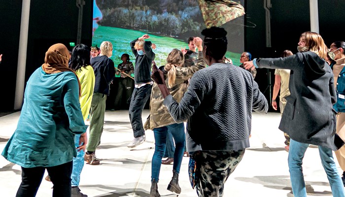 A large group of people are dancing in an industrial indoor space. They have their regular clothes on and are moving freeform. There is a large screen in the background showing a film with an outdoor scene and the profile of a person to the left.  Most of the dancers are facing the screen.