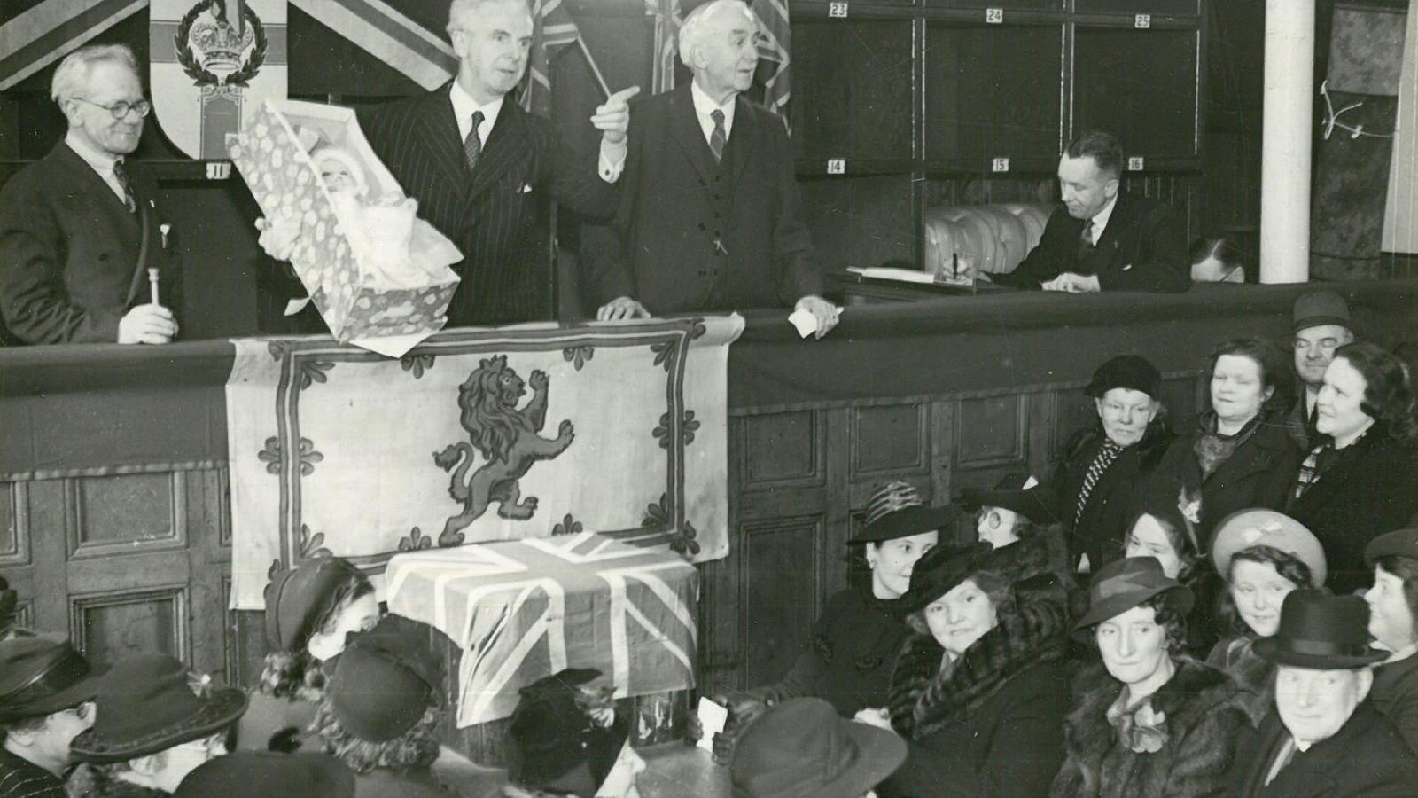 Three people standing talking to a crowd lower below, holding a baby in a small crib with a Lion Rampant of Scotland flag and a Scotland flag draped below.