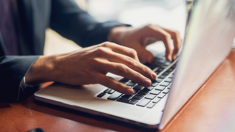 Close up a person's hands typing on a laptop. They are wearing a blue grey long sleeved top and you can see a glimpse of their watch.