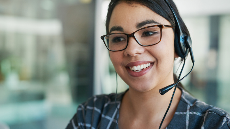 Person answering a phone with a headset on. They have glasses on and are wearing a checked dress.